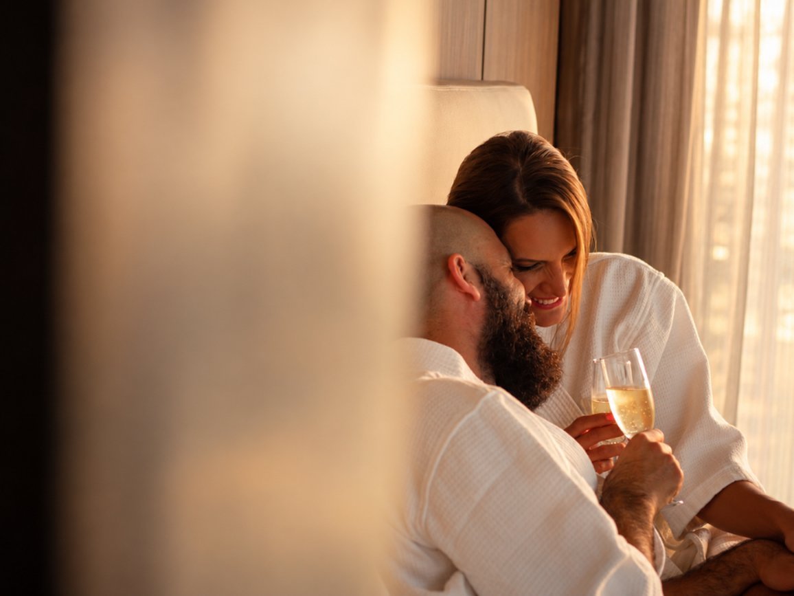 Pareja en albornoces blancos compartiendo una copa de espumante junto a una ventana